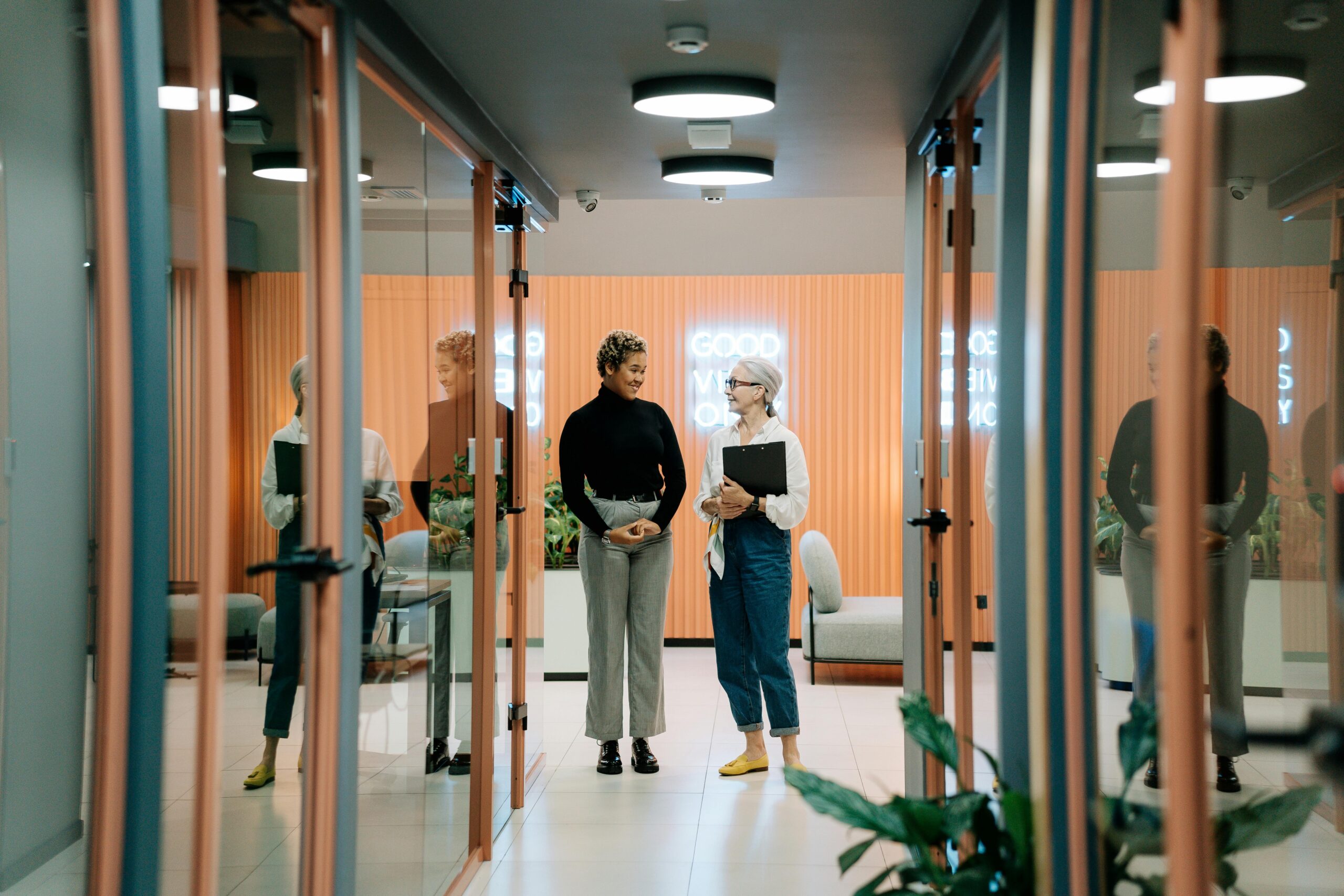 Two women discuss business in a stylish office hallway with glass doors and modern decor.